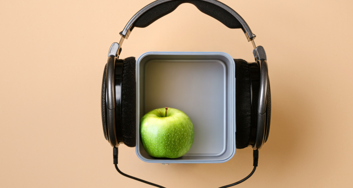A black pair of overhead headphones over a grey lunchbox with a green Granny Smith apple in it.